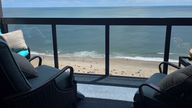 Ein Balkon mit Korbstühlen und Kissen, mit Blick auf einen Strand mit Menschen und Sonnenschirmen.