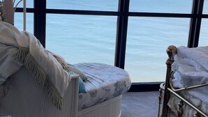 Ein Zimmer mit großem Fenster zum Meer, ein weißer möblierter Sessel und ein Bett mit Metallgestell.