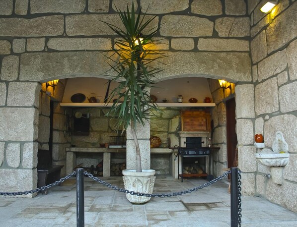 A stone-walled interior with a central potted plant, a fireplace, and shelves with various items.