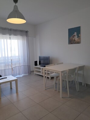 A living room with a TV, a dining table, chairs, a white sofa, and a painting on the wall. - TORRE 17- City View, Wifi, Cozy and Refurbished (Quarteira)