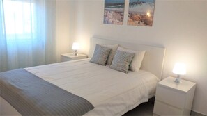 A neatly made bed with a grey throw, two bedside tables with lamps, and two wall-mounted artworks depicting beach scenes. - TORRE 17- City View, Wifi, Cozy and Refurbished (Quarteira)