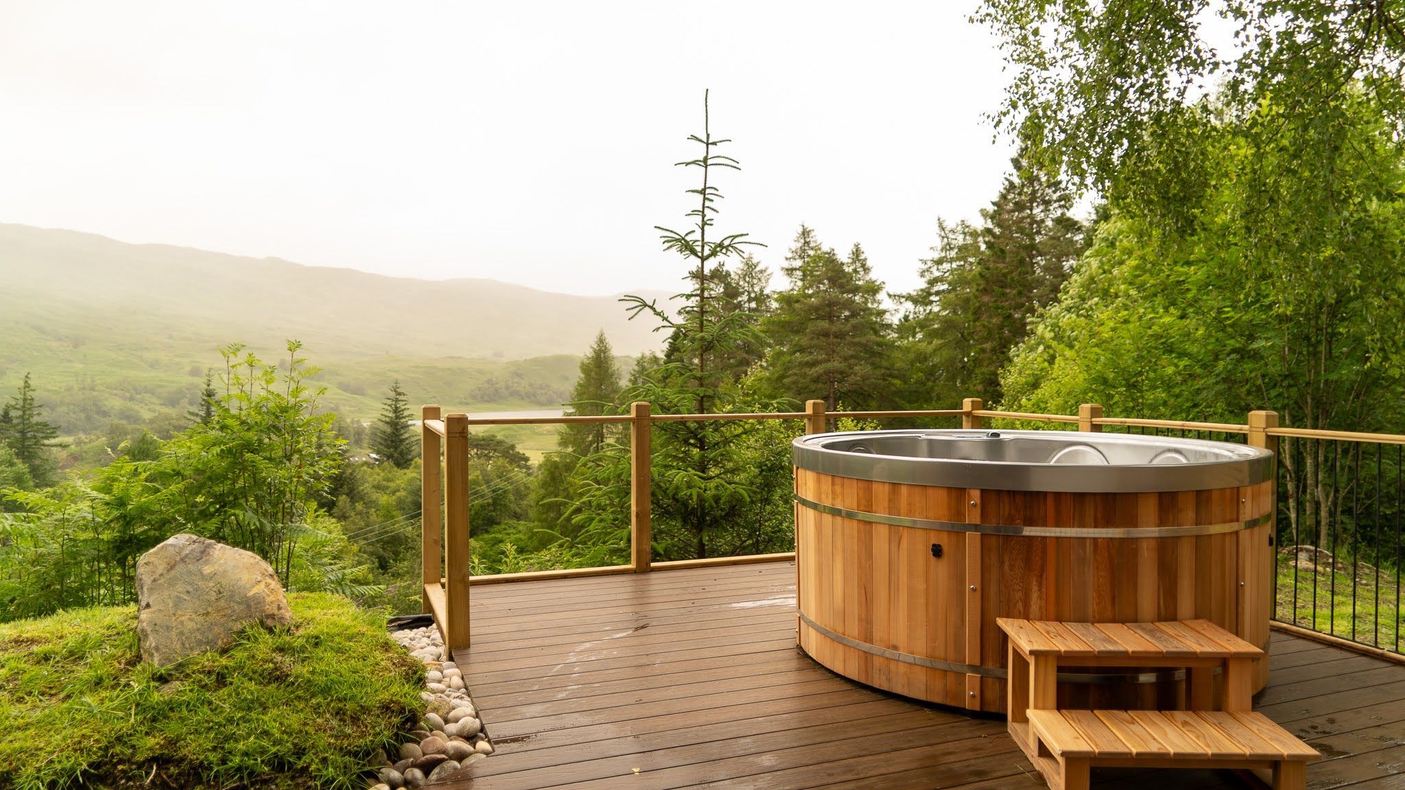 A hot tub on a wooden deck with a view of a forest and hills.