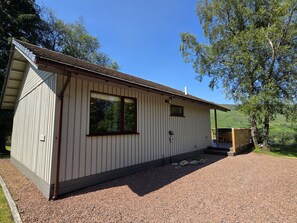 A single-story building with a sloped roof, a large window, and a gravel driveway. - Beautiful 4 star, 2 bedroom chalet with stunning loch and mountain views (Crianlarich)