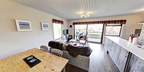 A cozy room with a sofa, a dining table, a kitchen area, and large windows. - Beautiful 4 star, 2 bedroom chalet with stunning loch and mountain views (Crianlarich)