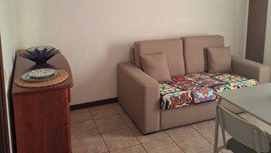 A living room with a beige sofa, a wooden side table, a patterned throw blanket, and a wall clock. - Beach House - 1st Floor (Moledo)