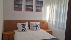 A bedroom with a bed, two wall-mounted pictures, a hanging light fixture, a nightstand, and a window with curtains. - Beach House - 1st Floor (Moledo)