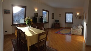 A dining area with a wooden table and chairs, a vase with flowers, and a view of mountains through large windows. - Panoramic apartment in Lake Como area (Asso)