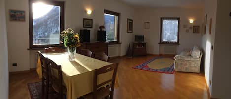 A dining area with a wooden table and chairs, a vase with flowers, and a view of mountains through large windows.