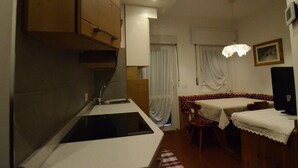 Cocina compacta con estufa integrada, lavamanos y armarios de madera. Hay una zona de comedor con dos mesas cubiertas con manteles de encaje y una silla.