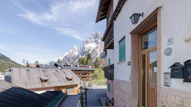 Un albergue de montaña con fachada de madera, entrada de piedra y vistas a picos nevados al fondo.