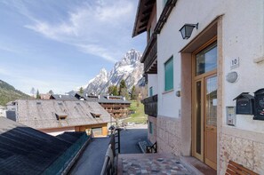 Un albergue de montaña con fachada de madera, entrada de piedra y vistas a picos nevados al fondo.