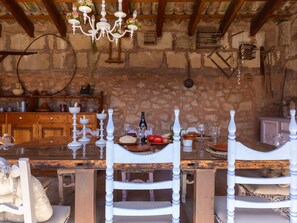 A rustic stone-walled room with a wooden dining table set for two, white chairs, and a chandelier.