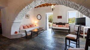A cozy living room with a stone floor, a wooden ceiling, and a large arched doorway. There is a sofa, a table, and a TV in the room. - Beachfront - Authentic- Idyllic- Relaxing -Homey - Sleeps 9 - Close to Naoussa  (Paros)