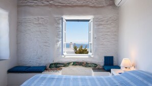 A bedroom with a bed, a window with a view of the sea, a small table with a lamp, and a blue pillow on the window sill. - Beachfront - Authentic- Idyllic- Relaxing -Homey - Sleeps 9 - Close to Naoussa  (Paros)