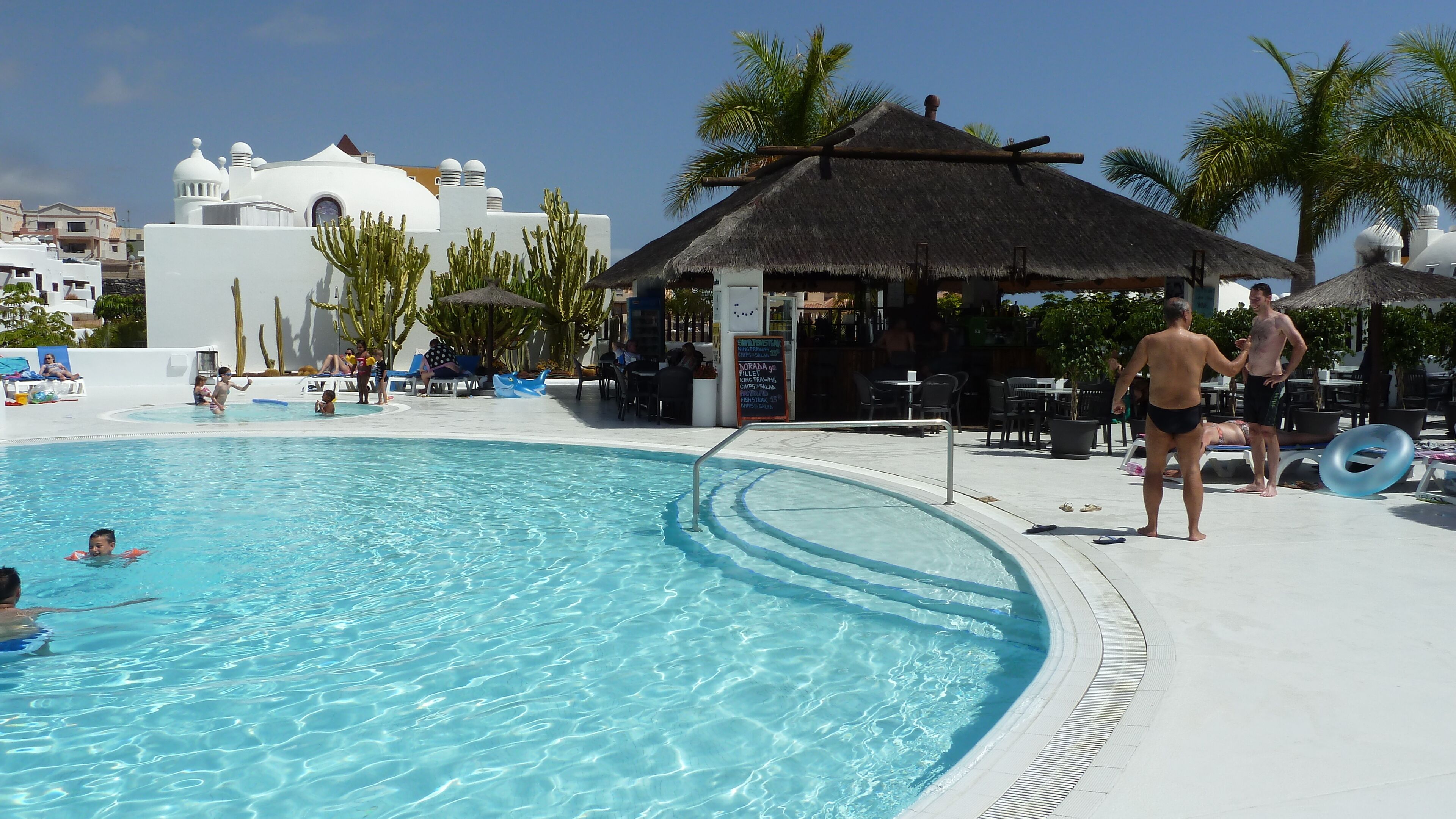 A pool area with a clear blue pool, lounge chairs, and a shaded area with a thatched roof.