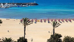 Una playa con una hilera de palmeras, un puerto deportivo con barcos y un faro al fondo.