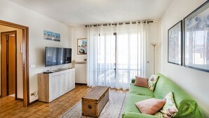 Uma sala de estar com um sofá verde, uma mesa de centro de madeira, uma televisão de tela plana e uma janela grande com cortinas leves.