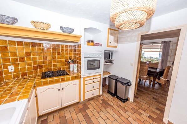 A compact kitchen with a gas stove, oven, and microwave, a small trash bin, and a wicker hanging light fixture. - House / Villa - LES ISSAMBRES (Roquebrune-Sur-Argens)