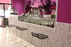 Uma área de bar com parede de pedra, dois banquetas pretas, divisória de vidro e prateleira com garrafas.