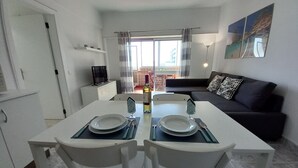 A dining area with a table set for two, a sofa, and a balcony with a view. - TORRE AZUL- Magnificent Sunset, Wifi, 20 m from the Beach (Quarteira)