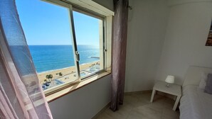 A bedroom with a large window overlooking a beach, a bed, a bedside table with a lamp, and a curtain. - TORRE AZUL- Magnificent Sunset, Wifi, 20 m from the Beach (Quarteira)