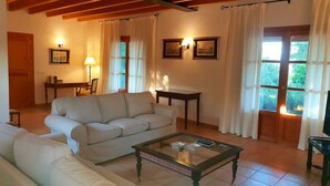 A living room with a sofa, a glass-top coffee table, and a wooden ceiling. There are two windows with curtains, a door, and framed pictures on the wall. - Majorcan finca in a paradise area with no neighbours  (Santa Margalida)