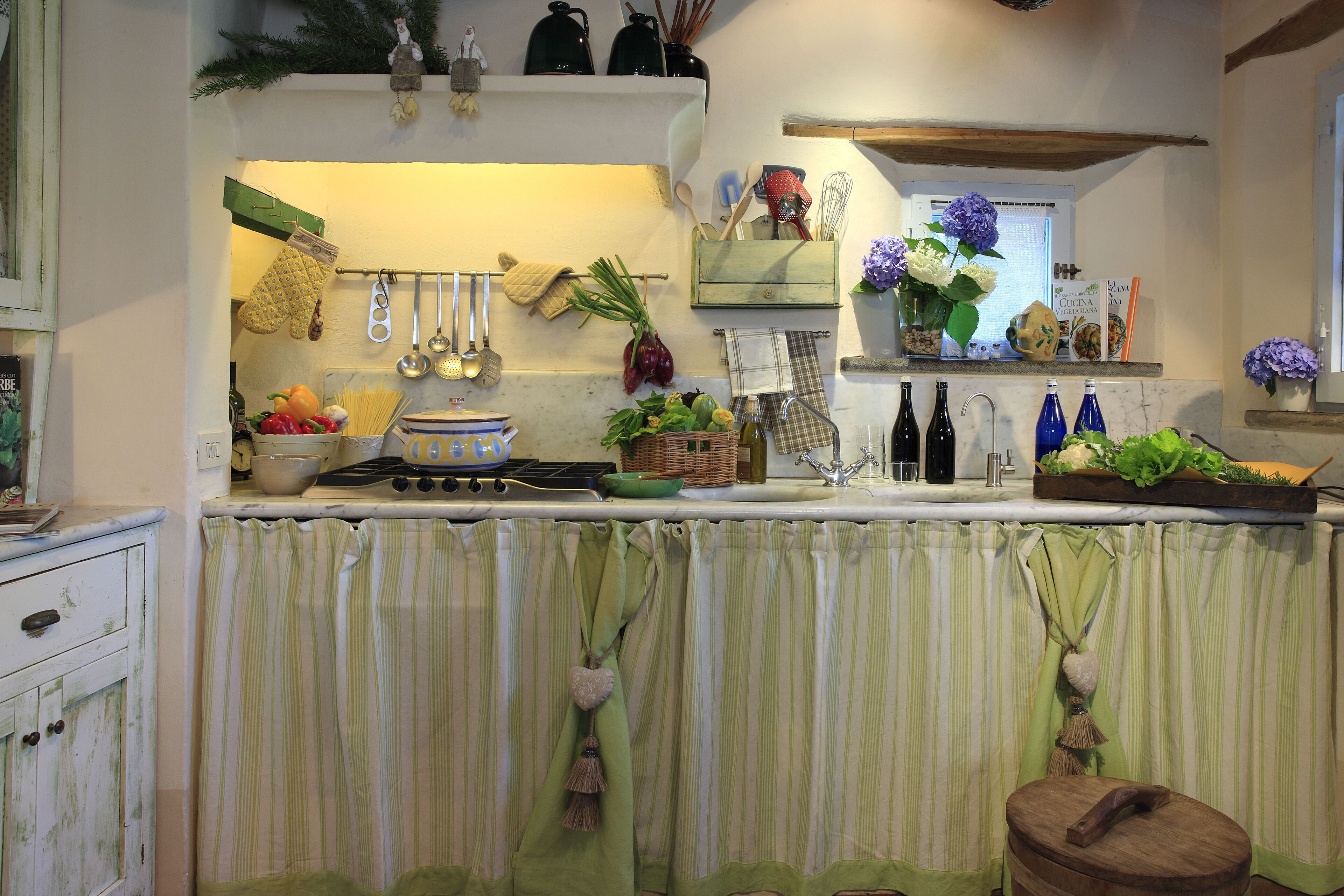 A kitchen with a stove, sink, and various kitchen utensils. There are hanging pots and pans, a cutting board, and a bowl of fruit.