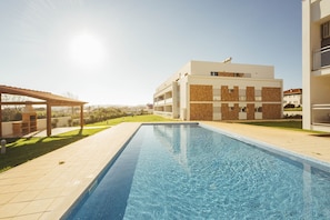 A swimming pool with a tiled deck, a covered barbecue area, and a modern building with balconies.