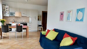 A modern kitchen with white cabinets, a dining table with chairs, and a blue sofa with colorful cushions.