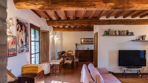Una sala de estar rústica con vigas de madera a la vista, una zona de comedor con mesa y sillas de madera, un sofá, un televisor y una estantería.