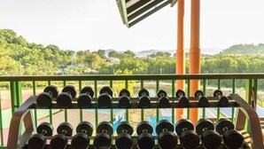 A gym with a row of dumbbells and a view of a green landscape with trees and a building in the distance. - T-House (Ratsada)