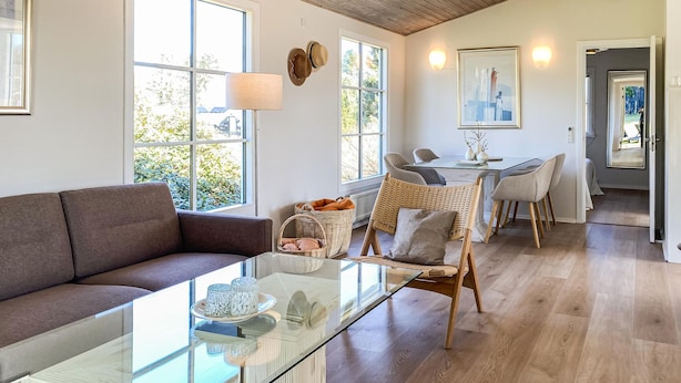 Een moderne woonkamer met een glazen salontafel, een bruine bank, rieten fauteuils en een eethoek met een tafel en stoelen.