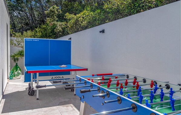 Una mesa de fútbol de mesa con tablero azul y figuras de jugadores rojo y azul, ubicada en un espacio exterior con pared blanca y follaje de fondo.