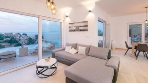 Uma sala de estar moderna com um sofá seccional cinza, uma mesa de centro redonda e vista para o mar através de grandes portas de vidro.