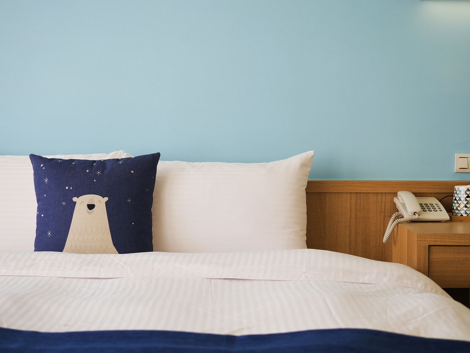 Una cama con almohadones blancos y azul marino, una mesita de noche de madera con un teléfono y una pared azul claro.
