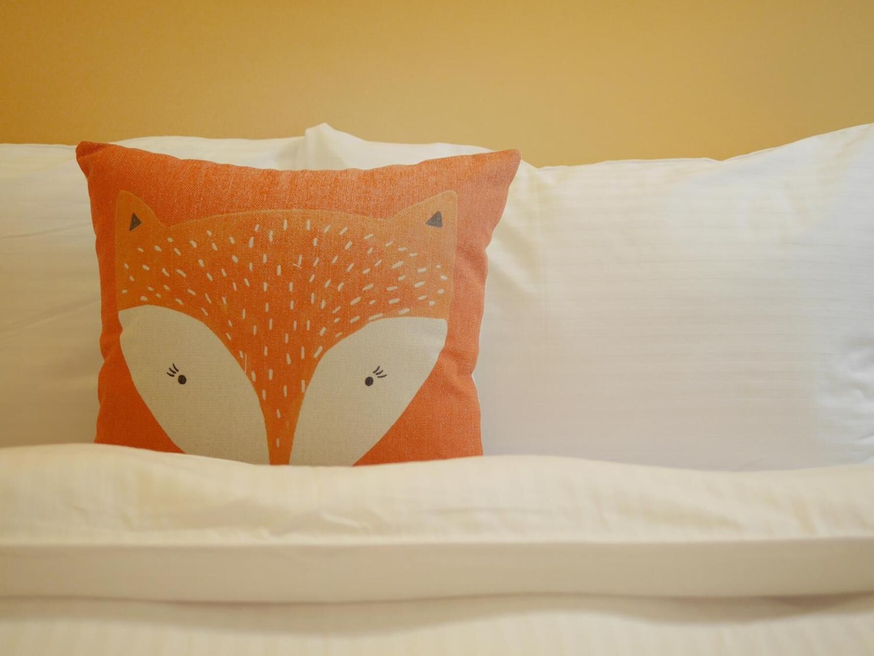 A bed with a white sheet and a single orange pillow with a fox design.