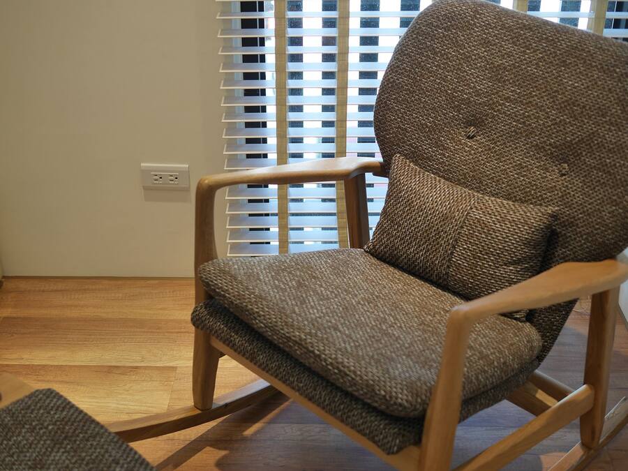 A wooden rocking chair with a woven fabric seat and backrest, positioned in a room with wooden flooring and a window with blinds.