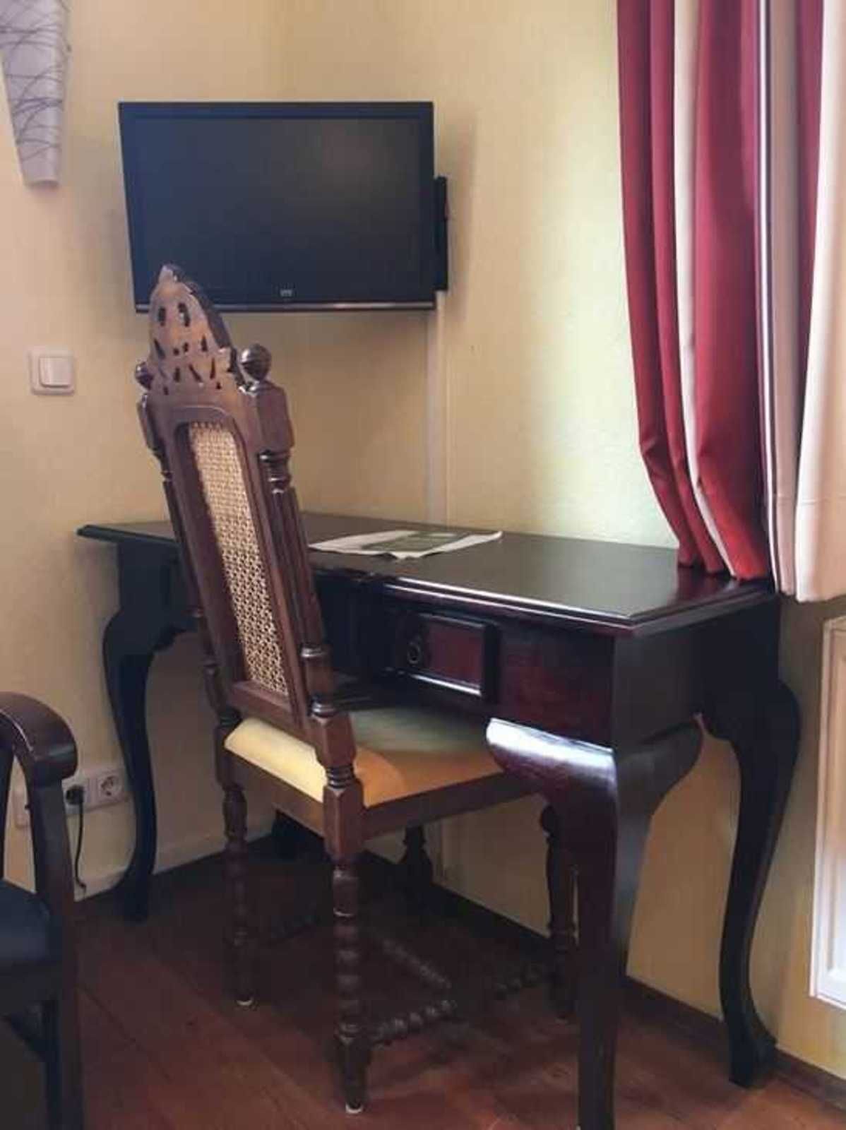 A room with a wooden desk, a chair, a television mounted on the wall, and a window with red curtains.