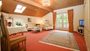 A cozy living room with a wooden ceiling, a large window with curtains, a television, and a red carpet. - Apartment 5, 77sqm, balcony, kitchen extra, 1 bedroom, 1 living room, 2 pers. (Ruhpolding)