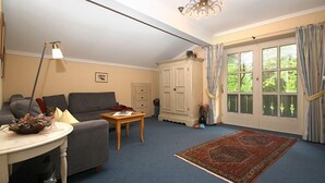 Ein Wohnzimmer mit einer Couch, einem kleinen Tisch, einer Lampe, einem Schrank und einem großen Fenster mit Vorhängen.