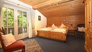 Ein gemütliches Schlafzimmer mit Holzbalkendecke, einem Doppelbett, einem Sessel und Blick ins Grüne durch das Fenster.