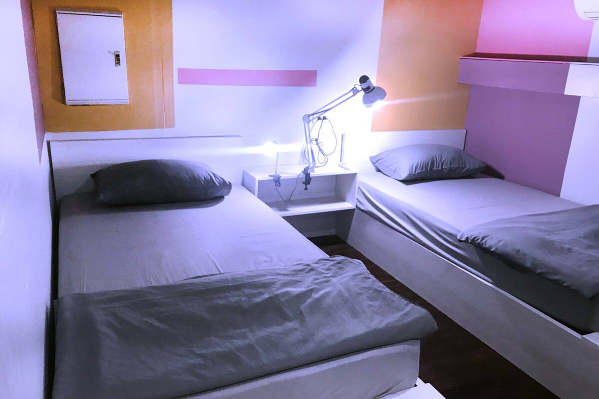 A twin room with two beds, a nightstand, a lamp, and a wall-mounted power outlet.