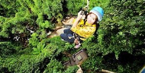 A person zip-lining through a lush, green forest with dense foliage and a small wooden platform at the end of the line. - Bed Bike Hostel (Chiang Mai)