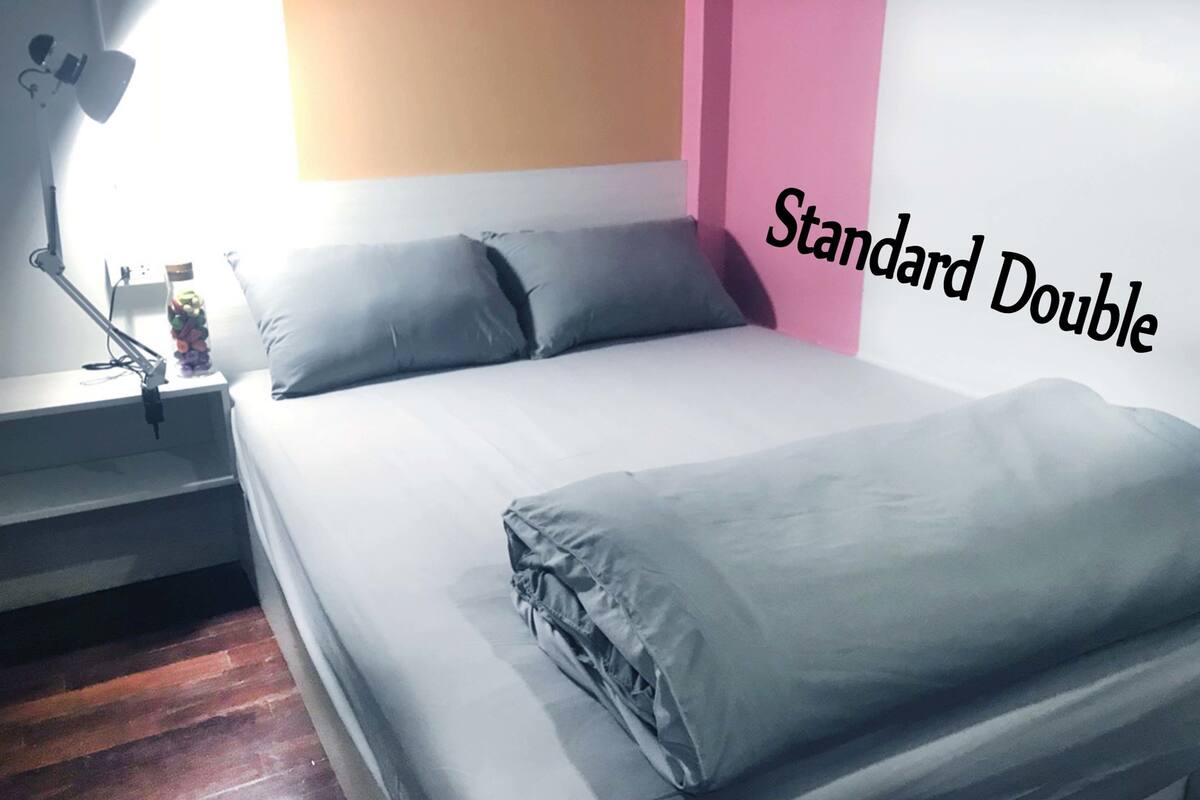 A standard double bed with grey bedding, a bedside lamp, and a pink wall.