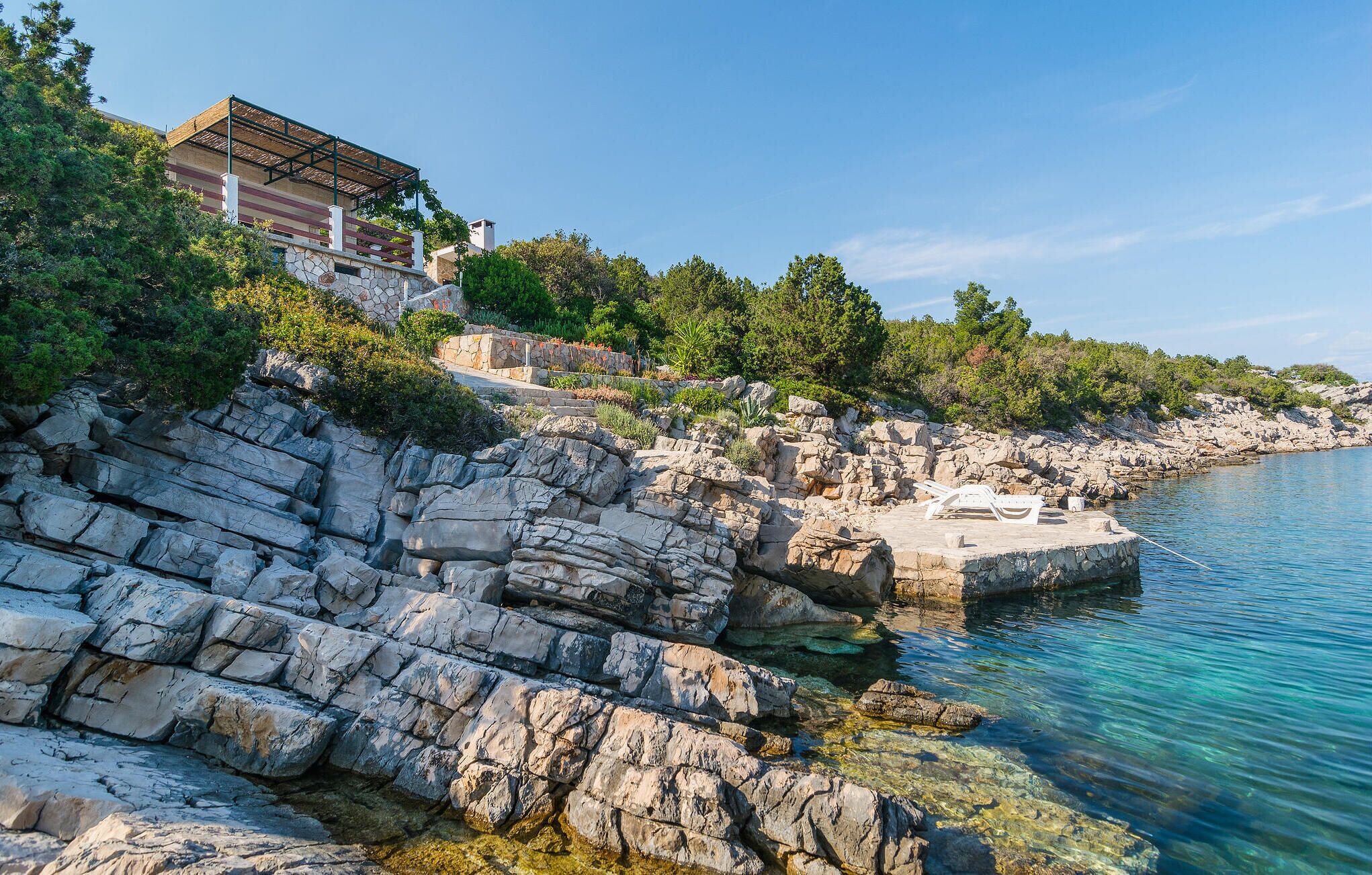 A coastal property with a stone facade, a wooden deck with lounge chairs, and a clear view of the sea.