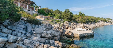 A coastal property with a stone facade, a wooden deck with lounge chairs, and a clear view of the sea.