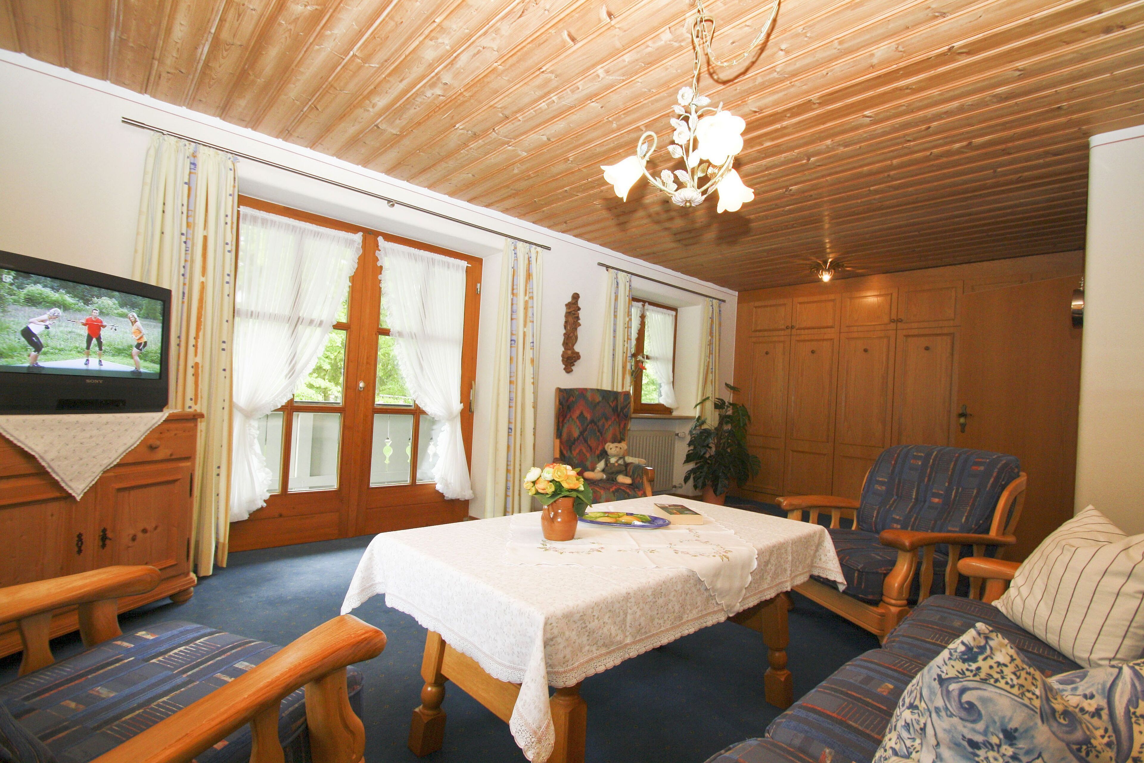 A cozy living room with a wooden ceiling, a television, a dining table with a white tablecloth, a blue sofa, and a wooden cabinet.