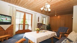 A cozy living room with a wooden ceiling, a television, a dining table with a white tablecloth, a blue sofa, and a wooden cabinet. - Apartment 2, 98sqm, balcony, kitchen, 2 bedrooms, 1 living room, max. 2 pers. (Ruhpolding)