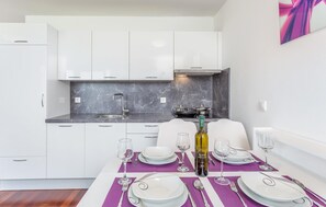 Una cocina moderna con gabinetes blancos, salpicadero de mármol y comedor con platos, cubiertos y copas de vino blancos.