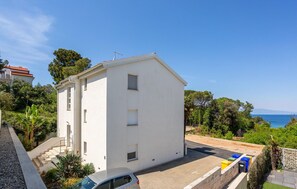 Un edificio blanco de dos pisos con un balcón, un coche aparcado delante y vista al mar.
